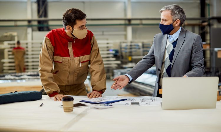 Mid adult businessman and male worker discussing about project plans while wearing protective face masks at carpentry workshop.